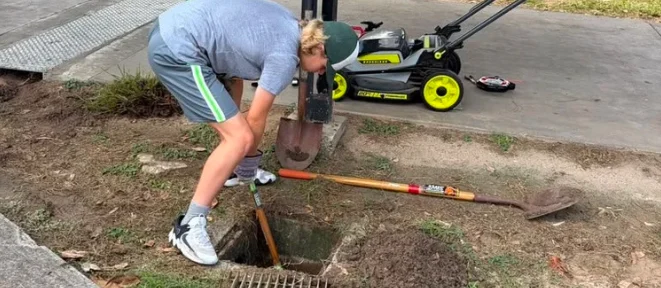 A student digging out a storm drain beside a campus sidewalk
